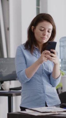Vertical video: Business worker browsing website on smartphone, using social media app in coworking space. Startup worker taking break from annual report planning with pc, holding mobile phone.