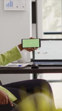 Vertical video: African american worker using horizontal greenscreen on mobile phone, looking at isolated copyspace display in business office. Young employee checking blank chroma key template on