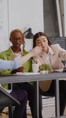 Vertical video: Diverse group of women doing teamwork to plan report, analyzing research statistics on papers in coworking space. Young employees attending briefing meeting to discuss investment.