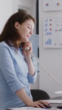 Vertical video: Business manager answering landline phone call in corporate space, working on company development. Professional assistant using office telephone with cord to have remote conversation.