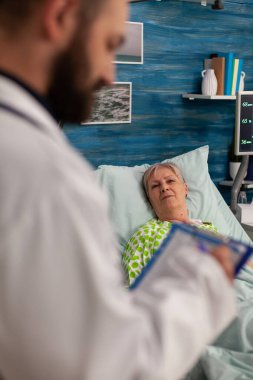 Retired woman lying in bed with chronic illness being cared for by medical specialist in nursing home room. Doctor taking notes on clipboard of elderly symptom of patient.