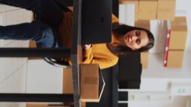 Vertical video: Young startup owner working on laptop to plan logistics, preparing products order to ship to clients. Female employee packing merchandise in boxes, taking notes for storage inventory.