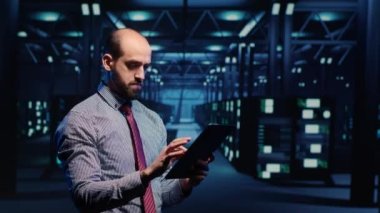 System administrator activating hardware in data center with tablet, analyzing modern servers in server room. Male infrastructure specialist looking at mainframe system. Tripod shot.