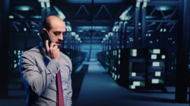Information security officer talking on phone call, working in professional data center with big data and server racks cabinets. IT technician using smartphone, inspecting network system. Tripod shot.