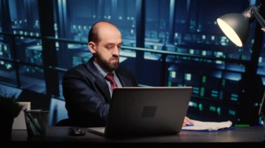 Male infrastructure engineer working in render farm running diagnostics on mainframe computer system in modern data center. Database admin controlling work of server cabinets on laptop.