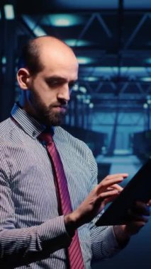 Vertical video: System administrator activating hardware in data center with tablet, analyzing modern servers in server room. Male infrastructure specialist looking at mainframe system. Tripod shot.