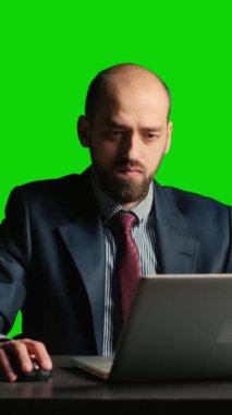 Vertical video: Entrepreneur looking at data on isolated greenscreen, and working on laptop. Young businessman in suit and tie standing over blank mockup and chroma key background, white collar.
