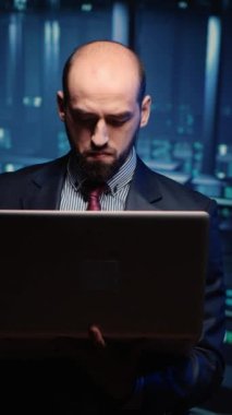 Dikey video: Sistem mühendisi dizüstü bilgisayarla sunucu kontrolü yapıyor, modern büyük veri merkezinde duruyor. Profesyonel ana bilgisayar donanımı üzerinde çalışan bilişim teknisyeni. Üçayak atışı.