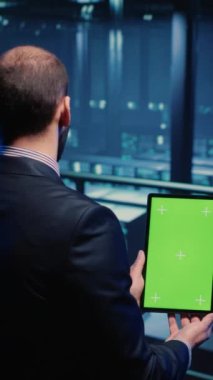 Vertical video: Data center technician holding tablet with green screen in server room, looking at isolated chroma key display. IT engineer inspecting mainframe computer processor, mockup template