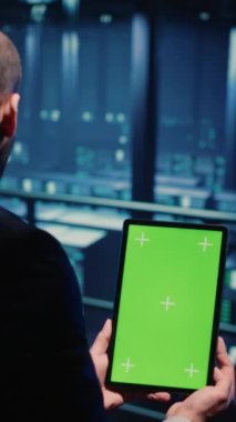 Vertical video: Network engineer doing system check on green screen, using tablet showing chroma key display with isolated copyspace in server room. IT technician working with cyber security. Tripod