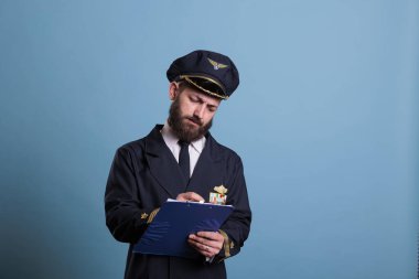 Airplane captain holding clipboard, concentrated pilot writing papers. Aircraft aviator in professional aviation uniform filling form in airport front view, studio medium shot on blue background