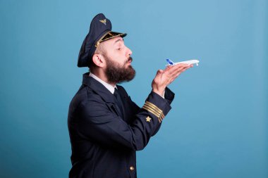 Airplane captain in uniform holding plane model in hands side view, pilot blowing at commercial passenger jet toy. Aircraft flying concept, professional aviator, studio medium shot