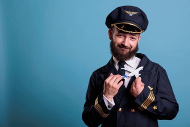 Smiling pilot in command wearing uniform holding small airplane model front view, aviation academy aviator playing with commercial plane toy. Aircraft captain looking at camera, studio medium shot