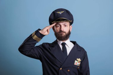Serious aviation academy airplane pilot saluting, wearing uniform and hat front view portrait, plane captain looking at camera. Middle age aviator with airline wings badge on jacket