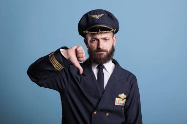 Serious airplane aviator with thumbs down gesture wearing professional uniform portrait, plane pilot looking at camera. Member aircrew showing disapproval sign with fingers, studio medium shot