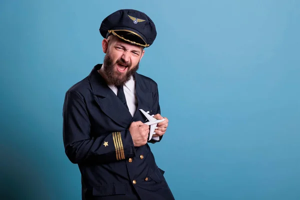 Funny pilot in uniform playing with small airplane model, aviation academy aviator holding commercial passenger plane toy. Aircraft crew member looking at camera, studio medium shot