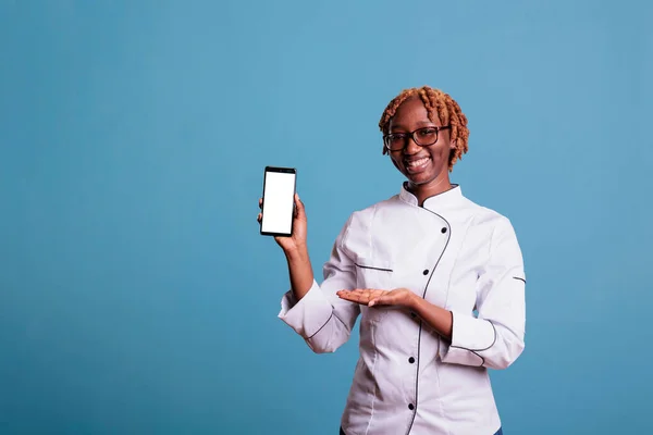 Female kitchen staff wearing work uniform holds cell phone with empty screen for advertising isolated on blue background. Happy african american cook smiling in studio shot.
