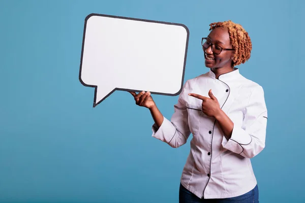 Smiling african american cook pointing to empty white dialogue bubble with copy space, showing advertising mockup. Cheerful kitchen worker with dialogue frame in studio shot.