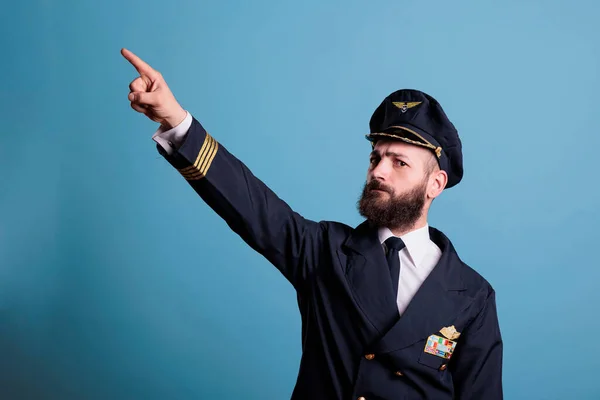 Serious captain pointing at sky with index finger, wearing professional aviation uniform, plane pilot looking at camera. Confident aviation academy aviator standing, studio medium shot