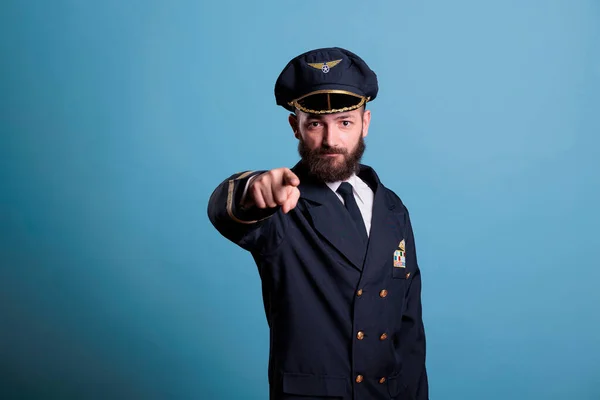 Serious airplane pilot pointing at camera, plane captain wearing uniform and hat front view portrait. Aviation academy aviator with airline wings badge on jacket looking at you
