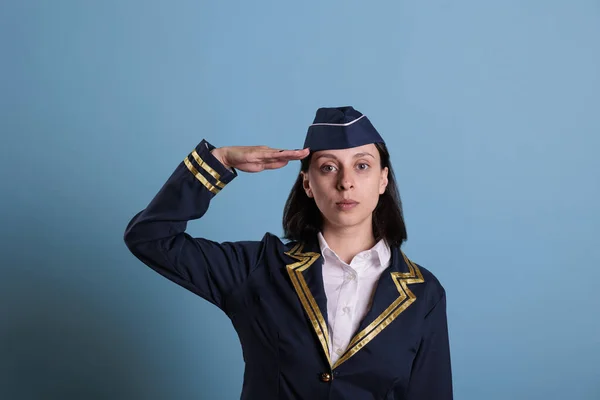 Portrait of flight attendant doing aviation honour salut in front of camera. Serious stewardess wearing professional flight uniform standing in studio with blue background, medium shot