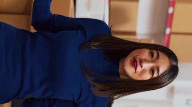 Vertical video: Young entrepreneur filming teleshopping video on phone, advertising products in warehouse space. Female worker using social media channel to create online ad promotion. Handheld shot.