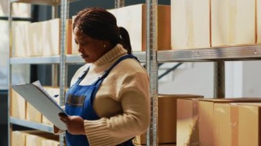 Young woman checking inventory list on clipboard, working with papers to see shipment order in storage room. African american person standing near shelves and racks in depot space.