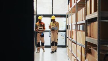Male workers using laptop and lists of logistics to plan stock inventory, looking at pallets of packs and boxes. Team of warehouse employees preparing supplies for distribution. Handheld shot.
