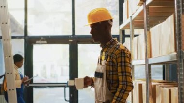 African american warehouse worker posing near shelves filled with products in boxes. Young man with overalls and helmet working in storage room depot with retail store merchandise.