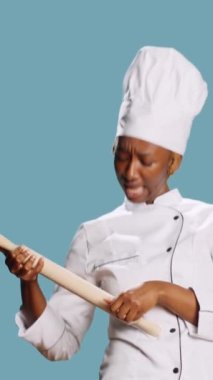 Vertical video: Cheerful funny chef playing with rolling pin and singing in studio, having fun with cooking tools. Young female cook holding wooden paddle gastronomy utensils over blue background.