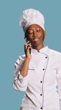 Vertical video: Professional chef talking on phone call in studio, having remote conversation using smartphone telephone line. Young person working as cook in culinary industry, answering mobile phone