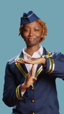 Vertical video: Young air hostess showing timeout symbol in studio, asking for break and rejecting work. Confident stewardess in flying uniform doing pause and refusal gesture over blue background.