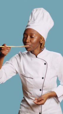 Vertical video: African american cook doing food taste test with spoon, sipping soup for taste testing on camera. Young gourmet chef in apron tasting restaurant meal over blue background, tasty food.