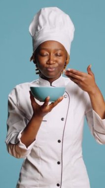 Vertical video: Cheerful gourmet chef smelling food aroma from bowl on camera, enjoying professional meal cooked. Professional female cook savoring tasty food, flavor and scent in studio. Catering