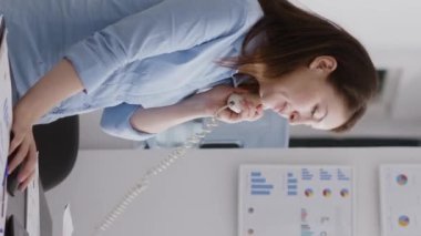 Vertical video: Business manager answering landline phone call in corporate space, working on company development. Professional assistant using office telephone with cord to have remote conversation.