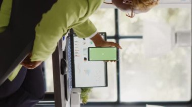 Vertical video: African american woman using greenscreen on phone, working with isolated chroma key display in startup office. Company employee checking blank mockup template on smartphone screen.