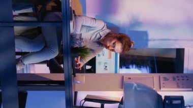 Vertical video: Young woman preparing to leave corporate job late at night, working after hours on financial growth report with marketing analytics. Business advisor leaving desk after finishing