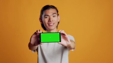Male model holding horizontal greenscreen mobile phone, showing isolated chroma key template on camera. Young person feeling happy using blank mockup display, checking copyspace screen.