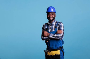 African american construction worker with happy face wearing a helmet smiling while looking at camera. Confident builder posing with arms crossed holding wrench against blue background.