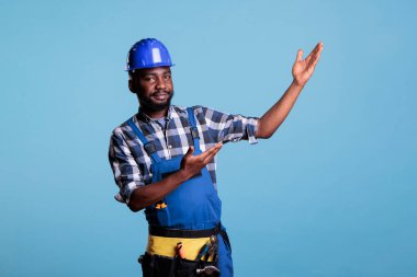 Construction worker raises hands to show an imaginary product, making advertising presentation with empty hands. Showing something as advertising, presenting gesture on blue background in studio.