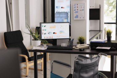 Nobody at company workstation desk with computer and tools, office wheelchair accessible. Empty workplace for business with annual data charts and research on monitor.