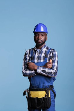 Portrait of construction professional in front of camera holding screwdriver in studio shot. African american builder wearing tool belt and work uniform isolated on blue background.
