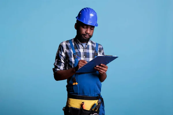 Construction supervisor writing on clipboard supervising building renovation work. Construction worker taking notes on sheet of paper to check job is perfect, studio shot against blue background.