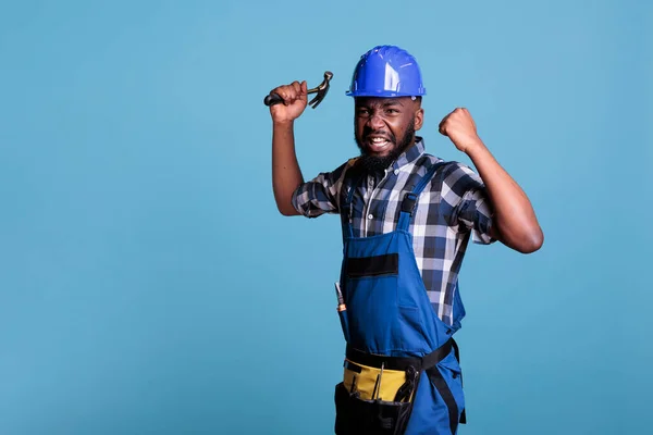 Frustrated employee feeling angry holding hammer and clenching fists in studio. Construction worker upset about working conditions showing teeth because of anger standing on blue background.