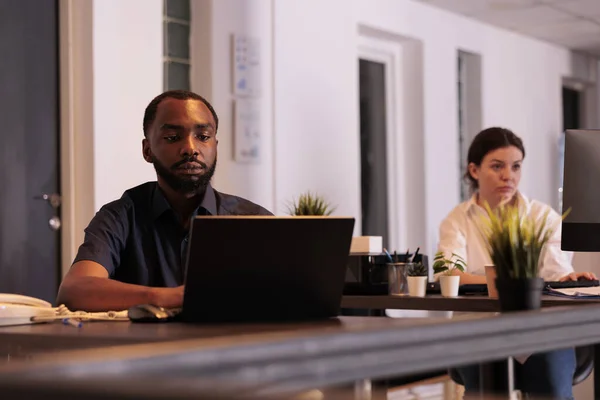 Employee analyzing report on laptop software in coworking space, project manager working late at office desk. African american man planning business strategy on computer at night