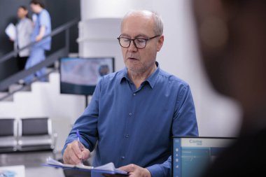 Elderly man holding clipboard signing medical documents before start consultation with doctor in hospital waiting area. Receptionist is managing patient records and scheduling follow-up appointments.