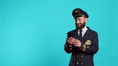 Uçuş ekibi kaptanı, stüdyoda internetteki akıllı telefonu kullanarak sosyal medya uygulamasını tarıyor. Mavi arkaplan üzerinde cep telefonu ve tarayıcıyla oynayan genç uçak pilotu.