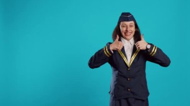 Young air hostess giving thumbs up in studio, showing okay like and approval gesture. Smiling stewardess in aviation uniform expressing agreement and success, doing good symbol on camera.