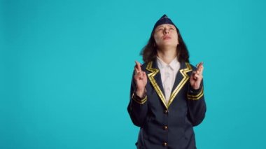 Positive air hostess standing with fingers crossed on camera, asking to fulfill wish over blue backdrop. Female aircrew member praying for luck and fortune, acting hopeful and wishful.