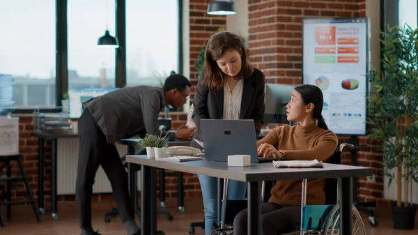 Woman with disability talking to colleague about business charts development to create strategy for financial growth. Office workers doing teamwork to plan project data on laptop.